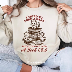 May include: A cream-colored sweatshirt with a graphic of three cats reading books. The text reads "I tried to form a gang but it turned into a book club" and "@MemoryArt666".