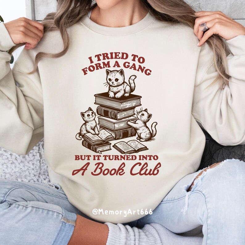 May include: A cream-colored sweatshirt with a graphic of three cats reading books. The text reads "I tried to form a gang but it turned into a book club" and "@MemoryArt666".