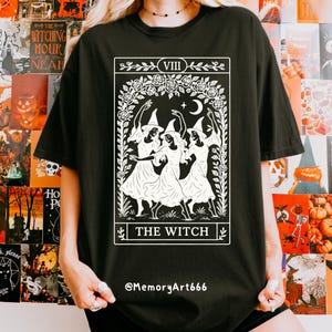 May include: Black t-shirt featuring a white graphic of three witches dancing under a crescent moon, framed by floral elements. The text "THE WITCH" is displayed below the image. The shirt also has the text "@MemoryArt666".