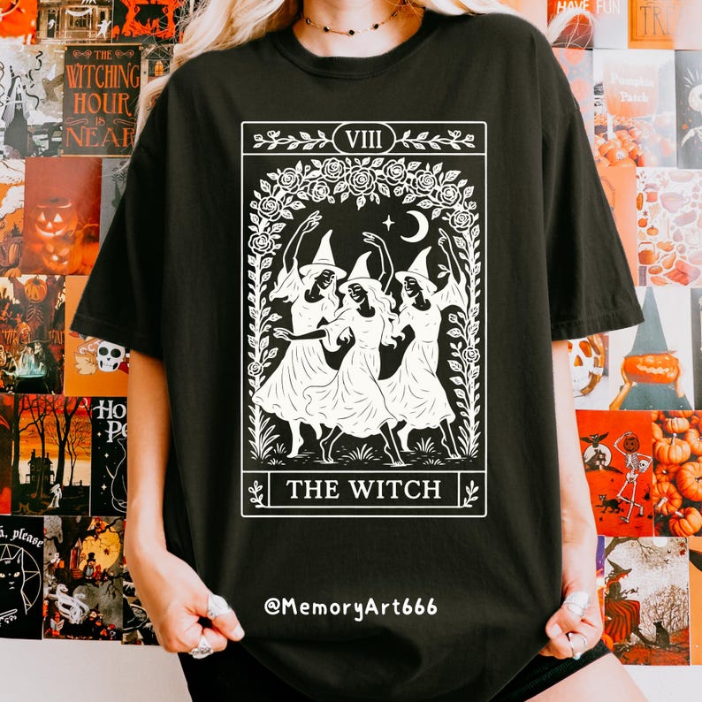 May include: Black t-shirt featuring a white graphic of three witches dancing under a crescent moon, framed by floral elements. The text "THE WITCH" is displayed below the image. The shirt also has the text "@MemoryArt666".
