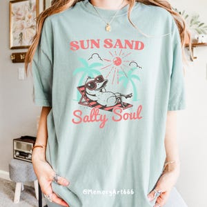 Peut inclure: Un t-shirt bleu clair avec un graphique d'un canard portant des lunettes de soleil et se relaxant sur une chaise de plage. Le texte "Sun Sand Salty Soul" est imprim&eacute; sur le t-shirt en rose.