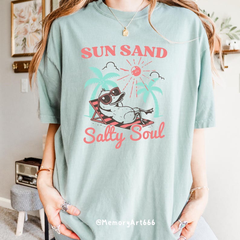 Peut inclure: Un t-shirt bleu clair avec un graphique d'un canard portant des lunettes de soleil et se relaxant sur une chaise de plage. Le texte "Sun Sand Salty Soul" est imprim&eacute; sur le t-shirt en rose.