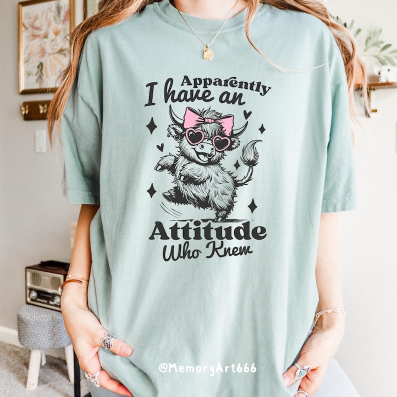 Op de afbeelding: Een lichtblauw t-shirt met een afbeelding van een Schotse hooglander die een roze zonnebril en een strik draagt. De tekst "Ik heb een houding, wie wist dat" staat op het shirt gedrukt.