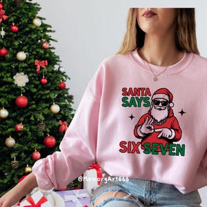 May include: A light pink sweatshirt with a graphic of Santa Claus wearing sunglasses and a red outfit. The text "SANTA SAYS SIX SEVEN" is printed on the front in green and red. A Christmas tree is in the background.