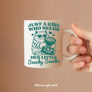 May include: White ceramic mug with a green design featuring a cartoon cat wearing sunglasses and holding a bag of snacks. The text on the mug reads "Just a girl who needs her little snacky snacks".