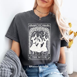 May include: Dark gray t-shirt featuring a white graphic of three witches dancing under a crescent moon and stars, framed by a floral wreath. The text "THE WITCH" is below the image. The shirt has short sleeves.