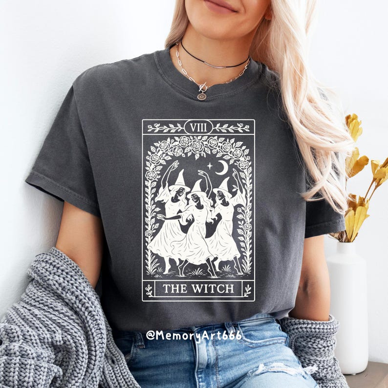 May include: Dark gray t-shirt featuring a white graphic of three witches dancing under a crescent moon and stars, framed by a floral wreath. The text "THE WITCH" is below the image. The shirt has short sleeves.