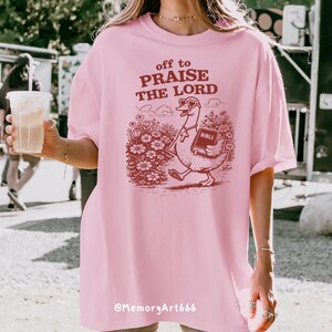 Op de afbeelding: Lichtroze T-shirt met de tekst "off to PRAISE THE LORD" en een cartoon gans met een bijbel. De gans loopt door een bloemenveld. De tekst "@MemoryArt666" staat onderaan het shirt.