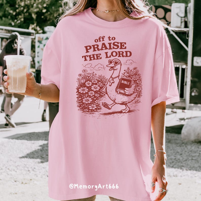 Op de afbeelding: Lichtroze T-shirt met de tekst "off to PRAISE THE LORD" en een cartoon gans met een bijbel. De gans loopt door een bloemenveld. De tekst "@MemoryArt666" staat onderaan het shirt.