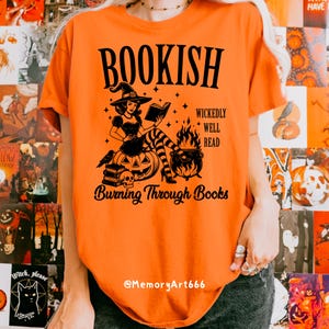 May include: Orange t-shirt with a black graphic featuring a witch reading a book, a cauldron, and the text "BOOKISH" and "Burning Through Books". The witch is sitting on a pumpkin. The shirt has a crew neck.