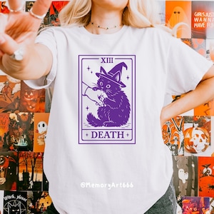 May include: White t-shirt featuring a purple tarot card design. The card depicts a black cat wearing a witch hat, with the word "DEATH" at the bottom. The text "XIII" is at the top. The artist's name, "@MemoryArt666", is at the bottom.
