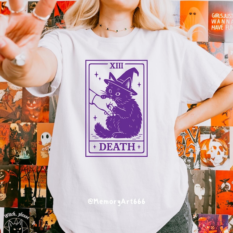 May include: White t-shirt featuring a purple tarot card design. The card depicts a black cat wearing a witch hat, with the word "DEATH" at the bottom. The text "XIII" is at the top. The artist's name, "@MemoryArt666", is at the bottom.