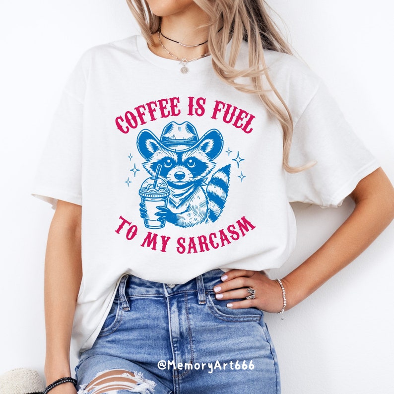 Puede incluir: Camiseta blanca con un mapache azul que lleva un sombrero de vaquero y sostiene una taza de caf&eacute;. El texto "COFFEE IS FUEL TO MY SARCASM" est&aacute; impreso en la camiseta en rosa.