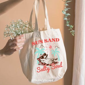 Peut inclure: Un sac fourre-tout en toile blanche avec un motif graphique rouge, bleu et vert. Le graphique repr&eacute;sente un canard de dessin anim&eacute; portant des lunettes de soleil et se relaxant sur une chaise de plage. Le texte "SUN SAND Salty Soul" est imprim&eacute; sur le sac.