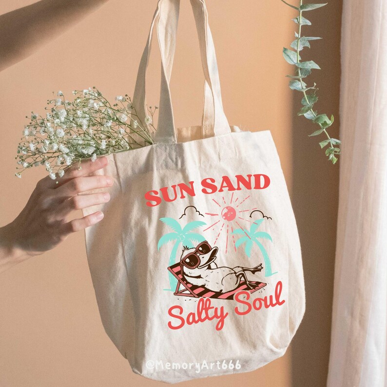 Peut inclure: Un sac fourre-tout en toile blanche avec un motif graphique rouge, bleu et vert. Le graphique repr&eacute;sente un canard de dessin anim&eacute; portant des lunettes de soleil et se relaxant sur une chaise de plage. Le texte "SUN SAND Salty Soul" est imprim&eacute; sur le sac.