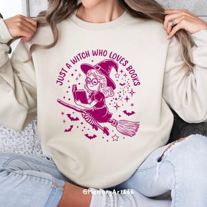 Może przedstawiać: Biała bluza z r&oacute;żowym nadrukiem przedstawiającym czarownicę czytającą książkę na miotle. Tekst głosi "Just a witch who loves books".