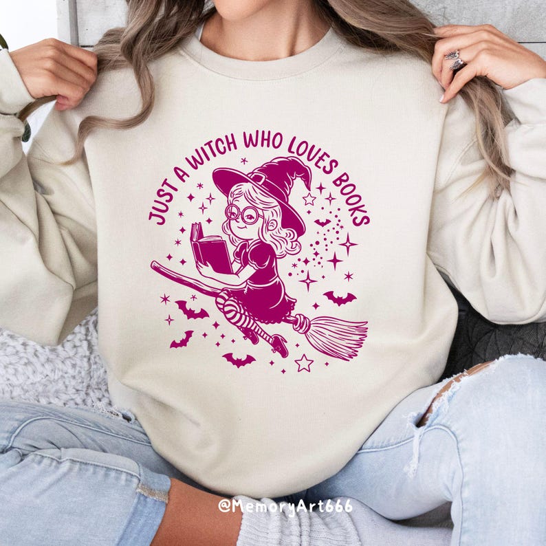 Może przedstawiać: Biała bluza z r&oacute;żowym nadrukiem przedstawiającym czarownicę czytającą książkę na miotle. Tekst głosi "Just a witch who loves books".