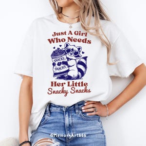 May include: White t-shirt featuring a graphic of a raccoon wearing sunglasses and holding a bag of snacks. The text "Just A Girl Who Needs Her Little Snacky Snacks" is printed on the shirt.