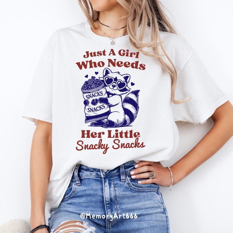 May include: White t-shirt featuring a graphic of a raccoon wearing sunglasses and holding a bag of snacks. The text "Just A Girl Who Needs Her Little Snacky Snacks" is printed on the shirt.