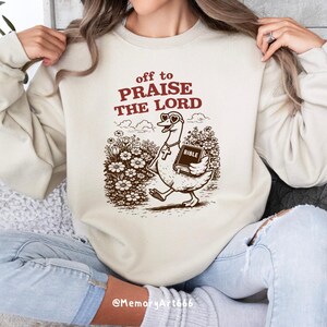 Op de afbeelding: Cr&egrave;mekleurige sweatshirt met de tekst "off to PRAISE THE LORD" in bordeauxrood. Een cartoon gans met hartvormige zonnebril en kruis ketting draagt een Bijbel. De afbeelding is in bruintinten.