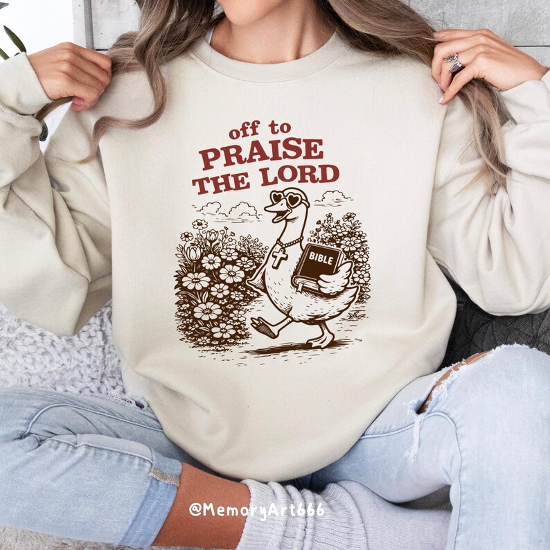Op de afbeelding: Cr&egrave;mekleurige sweatshirt met de tekst "off to PRAISE THE LORD" in bordeauxrood. Een cartoon gans met hartvormige zonnebril en kruis ketting draagt een Bijbel. De afbeelding is in bruintinten.