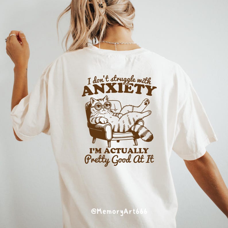 以下が含まれることがあります： 白いTシャツに、茶色の文字で「私は不安と闘っていません。実際、私はとても得意です。」と書かれています。このテキストには、眼鏡をかけた猫が椅子に座ってリラックスしているという漫画のイラストが添えられています。