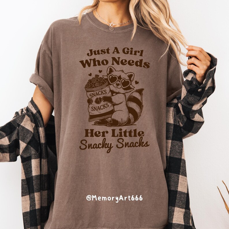 May include: A brown t-shirt with the text "Just a girl who needs her little snacky snacks" and a cartoon raccoon wearing sunglasses holding a bag of snacks. The text "@MemoryArt666" is at the bottom of the shirt.
