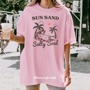 Peut inclure: T-shirt rose surdimensionn&eacute;e avec un graphique d'un canard de dessin anim&eacute; portant des lunettes de soleil et se relaxant sur une chaise de plage sous des palmiers. Le texte "Sun Sand Salty Soul" est imprim&eacute; sur le t-shirt. Le texte "@MemoryArt666" est imprim&eacute; en bas du t-shirt.