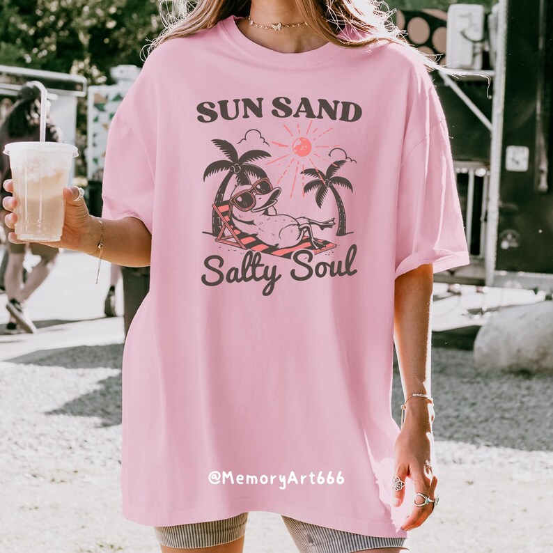 Peut inclure: T-shirt rose surdimensionn&eacute;e avec un graphique d'un canard de dessin anim&eacute; portant des lunettes de soleil et se relaxant sur une chaise de plage sous des palmiers. Le texte "Sun Sand Salty Soul" est imprim&eacute; sur le t-shirt. Le texte "@MemoryArt666" est imprim&eacute; en bas du t-shirt.