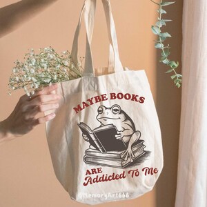 May include: A cream-colored tote bag with the text "MAYBE BOOKS ARE ADDICTED TO ME" and a graphic of a frog reading a book. The bag has long handles and is made of a canvas-like material. The text and graphic are in a dark red color.