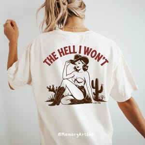 May include: White t-shirt featuring a brown illustration of a woman wearing a cowboy hat and boots. The text "The Hell I Won't" is printed in brown on the shirt.
