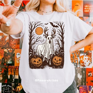 May include: White t-shirt featuring a Halloween-themed graphic. The design includes a ghost with deer antlers, a full moon, bare trees, pumpkins, and a bat. The text "@MemoryArt666" is at the bottom of the shirt.