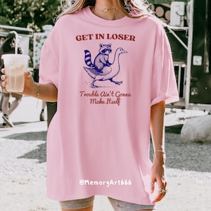 May include: Pink oversized t-shirt with a graphic of a raccoon riding a goose. The text on the shirt reads "Get in Loser, Trouble Ain't Gonna Make Itself" and "@MemoryArt666".