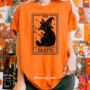May include: Orange t-shirt featuring a black graphic of a cat wearing a witch hat, with the text "XIII DEATH +" in a rectangular frame. The shirt has a crew neck and short sleeves. The text "@MemoryArt666" is at the bottom.