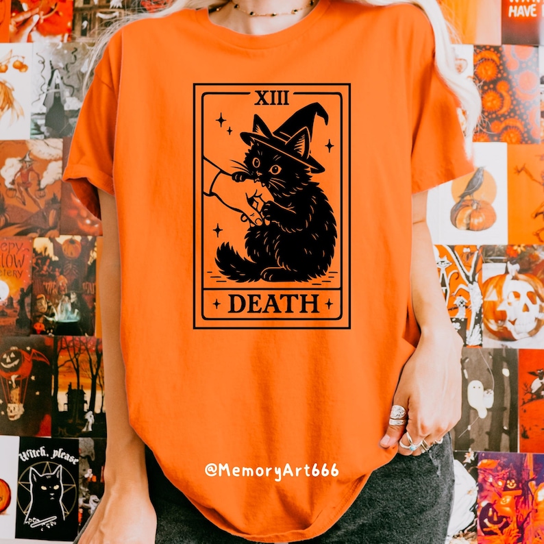 May include: Orange t-shirt featuring a black graphic of a cat wearing a witch hat, with the text "XIII DEATH +" in a rectangular frame. The shirt has a crew neck and short sleeves. The text "@MemoryArt666" is at the bottom.