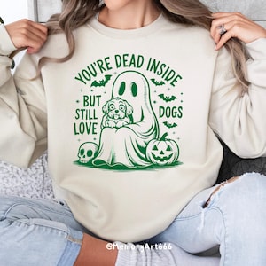 May include: A cream-colored sweatshirt with a green graphic of a ghost holding a dog. The text on the sweatshirt reads "You're Dead Inside But Still Love Dogs".