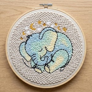 Peut inclure: Un cercle de broderie beige représentant un éléphant endormi. L'éléphant est dans des tons de bleu et de jaune, avec un contour noir. Des étoiles et des croissants de lune dorés sont brodés au-dessus de l'éléphant. Le cercle est posé sur un fond en bois clair.