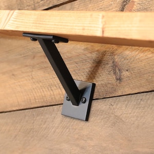 May include: Black metal shelf bracket with a square base and a single arm extending out to support a shelf. The bracket is mounted to a wooden wall.