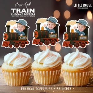Pode incluir: Três cupcakes cobertos com glacê branco, cada um adornado com um topper de cupcake de trem personalizado. Os toppers apresentam a imagem de uma criança com um chapéu de condutor, com "MASON EXPRESS". Os toppers medem 7,1 x 7,1 cm.