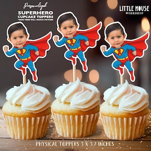May include: Three cupcakes topped with white frosting, each adorned with a superhero-themed topper. The toppers feature a child's face in a superhero costume with a red cape and blue suit. The toppers are 3 x 3.7 inches.