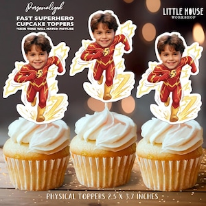 May include: Three cupcakes topped with white frosting and superhero-themed toppers. The toppers feature a child in a red and yellow superhero costume with the number 5. Text on the image reads "Personalized FAST SUPERHERO CUPCAKE TOPPERS" and "PHYSICAL TOPPERS 2.5 X 3.7 INCHES."