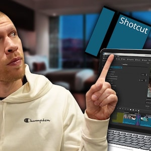 May include: A person points to a laptop screen showing the video editing software "Shotcut". The software interface is visible, with a video timeline and a preview window showing a landscape scene.