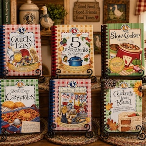 May include: Six spiral-bound Gooseberry Patch cookbooks displayed on black metal stands. Titles include Country Quick & Easy, 5 Ingredients or Less, Slow-Cooker Recipes, Best Ever Casseroles, Hometown Favorites, and Celebrate Year Round.