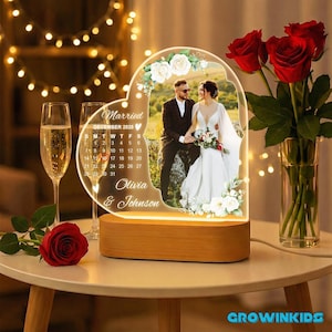 May include: A heart-shaped LED lamp with a photo of a couple, white floral accents, and the text "Married December 2025, Olivia & Johnson." The lamp sits on a wooden base. A bouquet of red roses and champagne glasses are in the background.