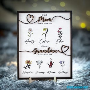 May include: A wooden sign with a heart design and the text "First - Mom Growing Since 1973" and "Now - Grandma Growing Since 2008". The sign also features flowers and names, including Amity, Celina, Edna, Danielia, January, Marian, and Anthony.