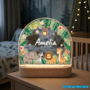 May include: A personalized night light with the name "Amelia" and the date "01-01-2026" displayed on a clear, arched panel. The panel features jungle-themed illustrations of animals, including an elephant, zebra, lion, giraffe, and monkey, set on a wooden base.