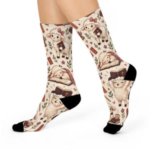 May include: Cream-coloured crew socks featuring a repeating pattern of cartoon lambs, open books with crosses, small flowers, and hearts. The lambs are depicted standing, sleeping on books, and holding books. The socks have solid black heels and toes.