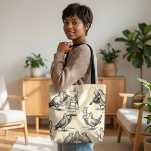 May include: Cream tote bag with black and white illustrations of doves as an artist, astronomer, and violinist. The bag has a black shoulder strap and is held by a person.