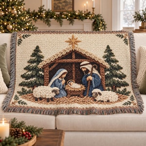 May include: A woven throw blanket featuring a nativity scene with a central stable, a golden star, and figures of Mary, Joseph, and the infant Jesus. The design includes two evergreen trees, two sheep, and a decorative fringed border edge.