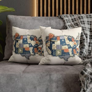 May include: Two white throw pillows with a patchwork design in the shape of the United States. The patchwork features blue, red, and white fabrics with floral and gingham patterns. The pillows are on a gray couch.